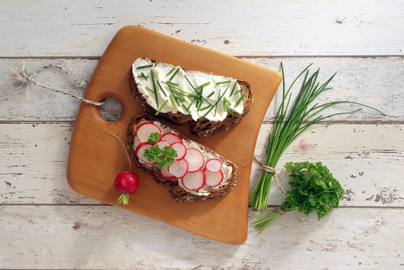 Due fette di pane di segale scuro guarnite con formaggio fresco biezpiens, erba cipollina fresca e ravanelli affettati su un tagliere di legno